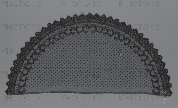 Veil, veils, lace, clothing accessories, fashion. Lace with a floral-geometric pattern running in a wide strip along the semicircular, serrated edge. The remaining part features fond de Tulle and regularly spaced small circles. 19th century, France. Clothing. Height 35 cm, width 74 cm., A semicircular piece of fabric laid flat against a gray background, primarily black in color with a sheer dotted mesh center and an ornate scalloped lace border; the lace border features floral and leaf motifs with a mix of solid and openwork patterns, and the mesh interior shows a regular arrangement of small dots; overall tones visible include black, dark gray, and the mid gray of the background, with a small tan tag near one edge.