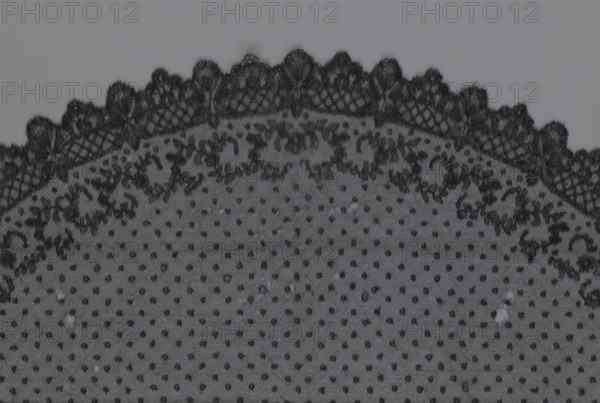 Veil, veil, lace, clothing accessory, fashion. Lace with a floral-geometric pattern running in a wide strip along the semicircular, serrated edge; remaining part features fond de Tull with regularly spaced small circles. 19th century, France. Clothing. Height 35 cm, width 74 cm., A close-up view of a piece of fabric with a delicate lace edge along the top, showing a scalloped border of intricate floral and leaf-like patterns in dark tones, transitioning into a dotted mesh field below; the visible colors are shades of gray and black, with fine netting texture, clusters of darker embroidered motifs, and small darker dots evenly dispersed across a lighter gray background.