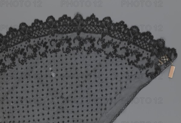 Veil, veils, lace, clothing accessories, fashion. Lace with a floral-geometric pattern running in a wide strip along the semicircular, serrated edge. The remaining part features fond de Tulle and regularly spaced small circles. 19th century, France. Clothing. Height 35 cm, width 74 cm., A section of black lace fabric with a scalloped floral edge and a dotted mesh body lying on a gray background, showing a few loose threads and a small beige rectangular tag attached near one corner.