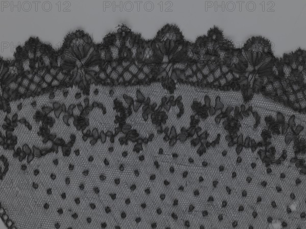 Veil, veils, lace, clothing accessories, fashion. Lace with a floral-geometric pattern running in a wide strip along the semicircular, serrated edge. The remaining part features fond de tulle and regularly spaced small circles. 19th century, France. Clothing. Height 35 cm, width 74 cm., A close-up of a piece of lace fabric showing a scalloped edge with ornate floral and leaf motifs, a netted background, and a dotted mesh area; the lace appears in shades of black and dark gray against a lighter gray backdrop.