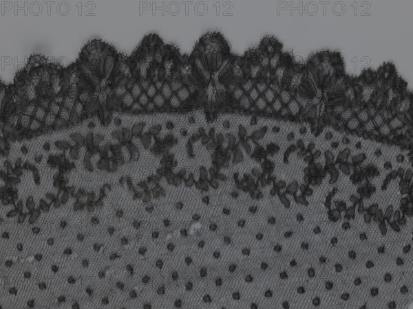 Veil, veils, lace, clothing accessories, fashion. Lace with a floral-geometric pattern running in a wide strip along the semicircular, serrated edge. The remaining part features fond de Tull and regularly spaced small circles. 19th century, France. Clothing. Height 35 cm, width 74 cm., A close-up view of a piece of black lace fabric with a scalloped top edge, featuring a floral and leafy embroidered pattern above a background of small raised dots on a fine hexagonal mesh; the visible colors are shades of black and gray.