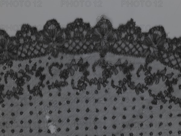 Veil, veils, lace, clothing accessories, fashion. Lace with a floral-geometric pattern running in a wide strip along the semicircular, serrated edge; the remaining part features fond de Tull and regularly spaced small circles. 19th century, France. Clothing. Height 35 cm, width 74 cm., A close-up view of a piece of fabric showing a scalloped lace edge with floral and leaf motifs, a mesh background, and a pattern of raised dots; the visible colors are shades of black and gray.