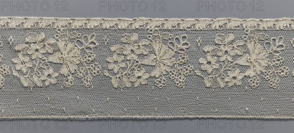 Lace fragment, Mid-19th century, France. Section of a straight strip used as a background for a floral point clair ornament. Edge is a narrow strip with a leaf motif. Above is a floral tendril composed by repeating bouquets, with a carnation cornflower? flower in the center. Next to the tendril are small dots in a regular pattern. Lace, width 10.5 cm, length 162 cm., A strip of delicate lace with a repeating floral pattern set against a gray background, the lace rendered in a cream or off-white color; the top edge shows a scalloped trim with small loops, below which a row of ornate flower and leaf motifs are connected by fine netting; the lower portion features a hexagonal mesh with occasional tiny raised knots, and the bottom edge is finished with a narrow stitched border; overall tones visible are cream or off-white and gray.