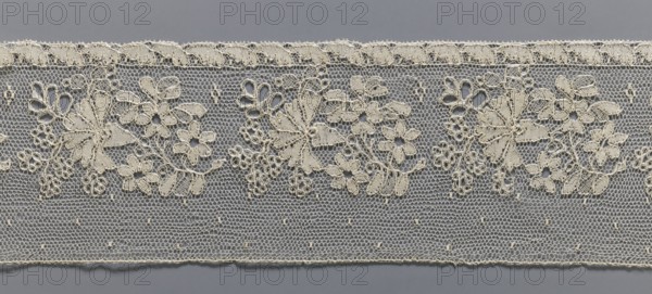 Lace fragment, As a background for the floral point clair ornament, the preserved fragment is a section of a straight strip. The edge is a narrow strip with a leaf motif. Above, a floral tendril composed by repeating a bouquet, with a carnation cornflower? in the center. Next to the tendril are small dots in a regular pattern. Mid-19th century, France. Lace, width 10.5 cm, length 162 cm., A horizontal band of delicate lace in pale cream is shown against a flat gray background, featuring a repeating floral design with a few larger stylized blossoms and several smaller flower clusters connected by looping stems and tiny circular motifs; the lace ground is made of a hexagonal open mesh with a scalloped cream edging along the top and a straight finished cream edge along the bottom, and the overall palette includes cream and gray.