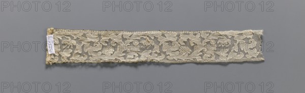 Two fragments of appliqués on batiste tulle: a with a floral tendril pattern; b with a fragment of cardboard sewn on. 19th century, Europe. Lace, embroidery and appliqué patterns; clothing accessory fabric. Width 5.5 cm, length 34 cm., A long narrow strip of lace with an ornate repeating floral and scrollwork pattern in a beige or off-white color is displayed on a flat gray background; the lace has a delicate net-like ground, slightly frayed edges, and a small fabric tag attached at one end, and the overall scene shows subtle shadows beneath the strip.