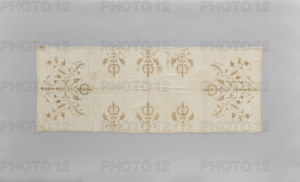 Church napkin of thin white linen 25 warp and weft threads per cm covered with embroidery featuring a pattern of large, evenly distributed, stylized Eastern flower bouquets. Flat, double-sided, pierced embroidery, using silk and gold thread. Very fine stitches. Cream and gold predominate in the ornament. Edged with cream veining. All edges with warp or weft threads ripped out to obtain a short fringe, approx. 1 cm long. 17th/18th century, Poland. Height 174 cm, width 65.5 cm. Subject: flowers, leaves, plant ornaments; sacred art; used as church paraments in Evangelical churches. Materials: linen, silk, gold thread., A rectangular fabric panel with a light beige background displayed flat against a pale gray backdrop, featuring symmetrical ornamental motifs in shades of brown and tan arranged across the surface; the cloth shows subtle wrinkles and a slightly frayed edge, with a few darker brown spots and stains visible, and several ornate floral and scroll-like designs positioned along t...