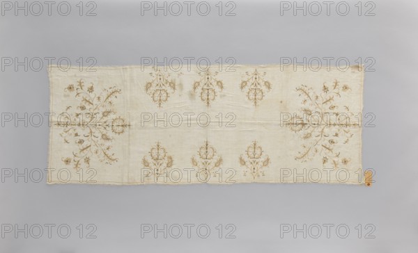 Church napkin of thin white linen 25 warp and weft threads per cm covered with embroidery featuring a pattern of large, evenly distributed, stylized Eastern flower bouquets. Flat, double-sided, pierced embroidery, using silk and gold thread. Very fine stitches. Cream and gold predominate in the ornament. Edged with cream veining. All edges with warp or weft threads ripped out to obtain a short fringe, approx. 1 cm long. 17th/18th century, Poland. Materials: linen, silk, gold thread. Height 174 cm, width 65.5 cm. Function: church napkin; motif: flowers, leaves, plant ornaments; sacred art; used in churches and chapels., A rectangular textile laid flat on a neutral background showing a pale cream or off-white base with decorative floral motifs in a muted tan or light brown. The fabric has frayed edges and visible creases and folds. Symmetrical ornate floral arrangements appear along the length, with a few larger, branching spray designs near the shorter ends and several smaller, repeatin...