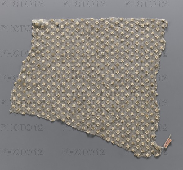 Lace fragment, lace, in a regular projective ornament with three motifs: a square, a quatrefoil, and four closely spaced circles. The preserved fragment has the form of a trapezoid and all edges are cut. 19th century, France. Height 38 cm, width 37 cm., A triangular piece of delicate net fabric laid flat on a solid background, with frayed uneven edges and a repeating embroidered motif of small floral-like shapes and diamond shapes across its surface; the fabric appears off-white or cream while the background is a cool mid-gray, and there is a small rectangular tag attached near one corner showing a hint of pale pink and beige.
