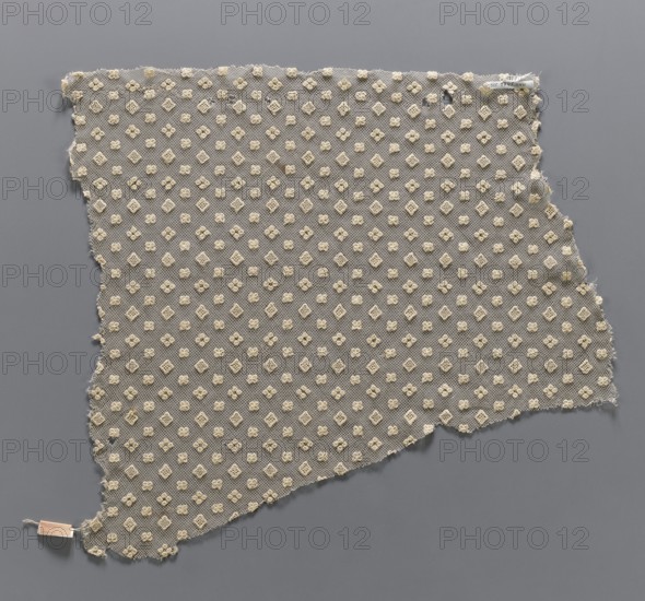 Lace fragment, in a regular projective ornament with three motifs: a square, a quatrefoil, and four closely spaced circles. The preserved fragment has the form of a trapezoid with all edges cut. 19th century, France. Lace; height 38 cm, width 37 cm., A torn piece of sheer fabric with a mesh-like gray background covered by a regular pattern of small raised floral and diamond motifs in cream, laid flat against a medium gray surface; frayed edges are visible around the irregular perimeter and a small beige tag is attached near one corner.