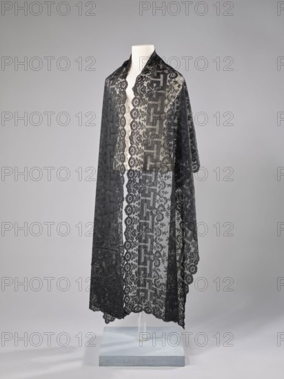 Shawl, shawl, fashion, women's clothing, clothing accessory, floral ornaments, meander ornament. The meander is associated with straight flower branches with a single large flower directed towards the outer edge. The outer edge is serrated. 1st half of the 19th century, France. Clothing, lace. Dimensions: length 270 cm, width 63.5 cm., A long sheer black lace shawl draped over a white mannequin bust, with an ornate repeating floral and geometric lace pattern visible through its translucent fabric; the shawl has scalloped edges and hangs down to a pale blue square display base mounted on a clear stand, set against a smooth light gray background.