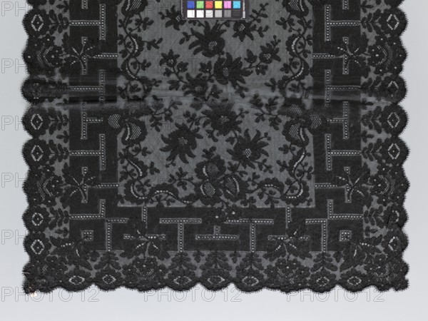 Shawl, shawl; shawls; fashion; women's clothing; clothing accessories; floral ornaments; meander ornament. The meander is associated with straight flower branches with a single large flower directed towards the outer edge. The outer edge is serrated. 1st half of the 19th century, France. Clothing, lace. Length 270 cm, width 63.5 cm., A rectangular piece of dark lace fabric with an ornate floral and geometric pattern occupies most of the image, showing a scalloped edge along the bottom and sides; the lace is primarily black with variations in shading where the intricate floral motifs, leaves, and vine-like scrolls contrast against a slightly lighter background, and a border of blocky geometric shapes alternates with floral elements around the interior; a small color calibration chart with several colored squares appears near the top edge, and the overall background behind the lace is a pale gray.