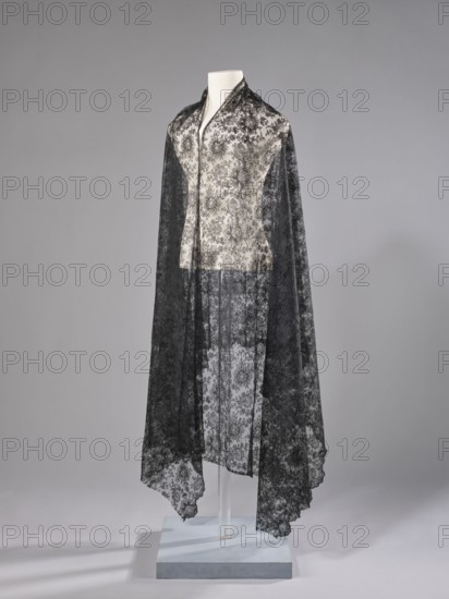 Shawl, shawl, floral ornaments, clothing accessory for women, fashion. Pattern on a mesh background. Long edges: upper edge sewn-on narrow stripe made using the bobbin technique; lower edge serrated without distinctive ornament. Short sides cut off and left unfinished. 1st half of the 19th century, France, fabrics. Dimensions: length 245 cm, width 100 cm., A mannequin draped with a long, sheer black lace shawl over a light-colored sleeveless top with a floral lace pattern; the shawl falls to the floor and has scalloped edges, creating a layered translucent effect that reveals the floral motifs beneath. The mannequin is displayed against a neutral gray background and stands on a pale rectangular platform with a clear vertical support visible beneath the garment. The visible colors are black, light cream or off-white, gray, and pale blue on the platform.