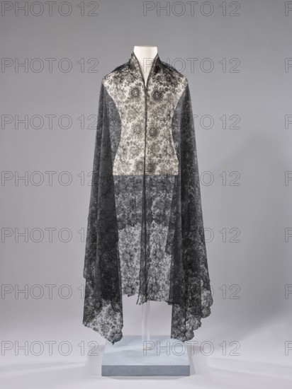 Shawl, shawl; floral ornaments; clothing accessory; women's clothing; fashion; pattern on a mesh background; long edges: upper — sewn-on narrow stripe made using the bobbin technique, lower serrated, without distinctive edge ornament; short sides cut off and left unfinished; 1st half of the 19th century, France : fabrics; length 245 cm; width 100 cm., A mannequin draped in a long, sheer lace cape with a floral pattern, showing a contrast between lighter lace at the upper front panel and darker lace panels along the sides and lower portion; the lace appears delicate and semi-transparent with scalloped edges at the hem, and the garment hangs down to a display stand that is pale and neutral in color; the background is a smooth, muted gray.