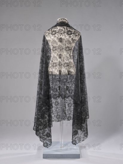 Shawl, shawl, floral ornaments, clothing accessory, women's fashion. Pattern on a mesh background. Long edges: upper — sewn-on narrow stripe made using the bobbin technique; lower serrated, without distinctive edge ornament. Short sides cut off and left unfinished. 1st half of the 19th century, possibly France. Fabric. Length 245 cm, width 100 cm., A mannequin draped with a long, sheer lace cape featuring a floral pattern in black over a light-colored background, the lace forming a darker overlay that extends down both sides and hangs to varying lengths with scalloped edges, displayed against a smooth gray backdrop and supported by a clear stand on a pale rectangular base.