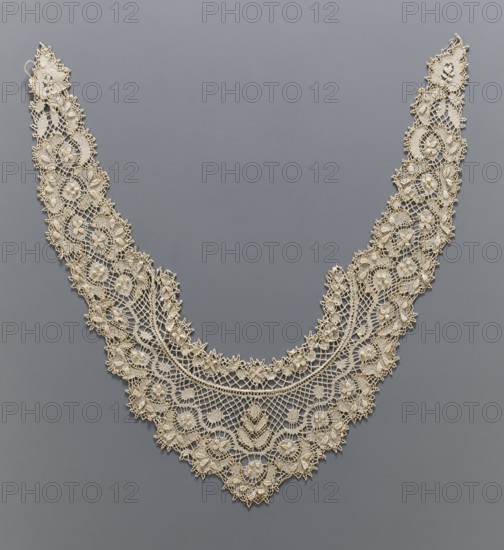 Collar, lace, ornamented with rosettes, floral leaves, and diagonal spider-like meshes arranged symmetrically. Horseshoe-shaped band with strongly elongated, pointed ends. 19th/20th century, Europe. Lace; length 96 cm, width 14 cm., Classification: collar; clothing accessories; women's clothing; fashion., A delicate V-shaped lace collar displayed against a smooth gray background, the lace appearing in an off-white or cream color with intricate floral motifs and geometric openwork; the design includes a scalloped outer edge, several rounded flower forms, leaf-like shapes, and densely stitched raised areas creating texture, with fine connecting threads forming mesh patterns and ornate clusters along the inner and outer curves.