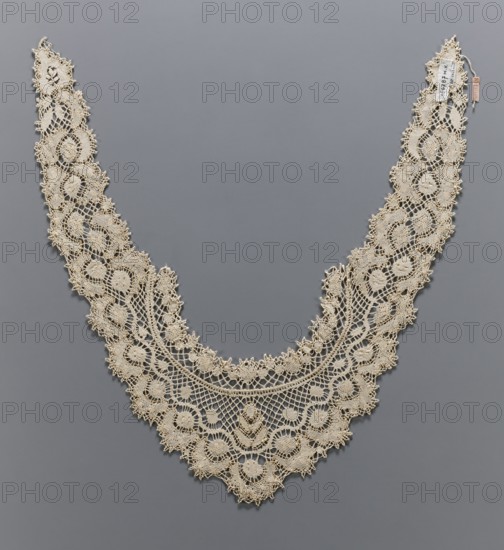 Collar, lace, collars, unknown origin, clothing accessory for women's fashion. Ornament with a pattern of rosettes and floral leaves and diagonal spider-like meshes; ornament composed symmetrically. The collar has the form of a horseshoe-shaped band with strongly elongated and pointed ends. 19th/20th century, Europe. Lace; length 96 cm, width 14 cm., A delicate cream-colored lace collar with an intricate openwork pattern is laid out against a smooth gray background; the lace forms a wide V shape with scalloped edges and repeating circular and floral motifs, showing fine netted sections and denser embroidered areas, and there are a few small tags attached near one end.