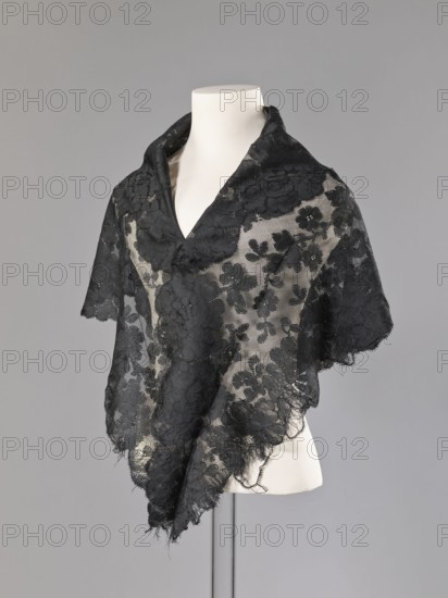 Cape, capes, fashion accessories, fashion, women's clothing. Cape in the form of an elongated rhombus, made of black lace with floral motifs. The edge is serrated in accordance with the forms of floral rosettes placed in a row along the edge. 2nd half of the 19th century, France. Garment, height 75 cm, width 164 cm., A dress form draped with a delicate black lace shawl featuring a floral pattern; the lace is sheer in places revealing the pale cream color of the form beneath, and the edges of the shawl are slightly frayed with a scalloped finish. The background is a smooth gray. The visible colors are black, cream, and gray.