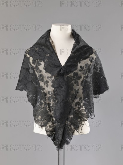 Cape in the form of an elongated rhombus, made of black lace with floral motifs. The edge is serrated in accordance with the forms of floral rosettes placed in a row along the edge. 2nd half of the 19th century, France. Garment; height 75 cm, width 164 cm., A dress form wearing a sheer black lace triangular shawl draped over the shoulders, with floral lace motifs and scalloped edges, the lace appearing delicate and slightly frayed in places, set against a plain light gray background; visible colors include black, off-white from the dress form, and shades of gray in the background.