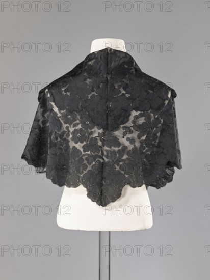 Cape, capes, fashion, women's clothing. Cape in the form of an elongated rhombus, made of black lace with floral motifs. The edge is serrated in accordance with the forms of floral rosettes placed in a row along the edge. 2nd half of the 19th century, France. Garment. Height 75 cm, width 164 cm., A black lace shawl with a floral pattern draped over a white mannequin torso against a neutral gray background, showing scalloped edges and sheer areas where the mannequin is visible; colors visible include black, white, and shades of gray.