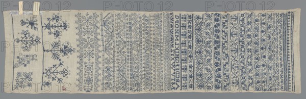 Embroidery sampler, Sampler with a cross-stitch pattern of trails, flower bushes, and letters. Blue embroidery on white canvas. Trails of geometric and floral ornaments with geometrized twigs and leaves. On one edge are several larger motifs in the form of stylized flower bushes. In the corner, the silhouette of a double-headed eagle. Between the paths, next to geometrical flower tendrils of various types, there is a double Holbein stitch ornament. 17th century, Silesia. Embroidery on fabric; height 81.5 cm, width 23 cm., A long rectangular piece of light-colored woven fabric embroidered with a dense array of blue motifs organized in vertical bands, with a few larger decorative floral and geometric motifs near one end and a group of narrow patterned stripes filling the rest. The embroidery shows intricate repetitive designs including floral, star-like, and angular shapes in varying shades of blue, with occasional small areas of darker blue contrast and a small isolated area of red near...