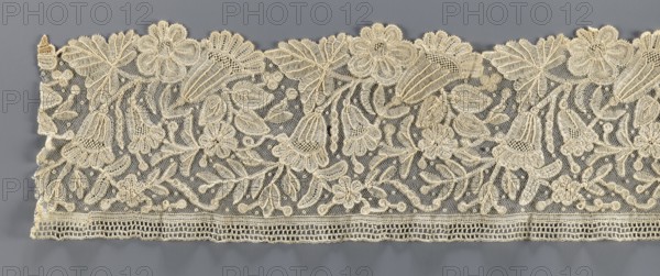Lace fragment, bobbin lace with relief edges made with a needle on a tulle background, featuring a motif of large flowers rosettes and bells and leaves. 19th century, Brussels. Lace, fabric; width 15 cm, length 110 cm., A horizontal length of lace with an off-white to cream color sits against a gray background, showing an openwork net ground decorated with a floral and leaf pattern in raised corded stitching; large stylized flowers, bell-shaped blossoms, clusters of small round buds, and elongated leaves form an overall repeating design, with scalloped upper edges and a straight lower edge featuring a narrow openwork border; the lace texture varies between fine netting and denser embroidered areas, and the visible colors are cream, off-white, and gray from the background.