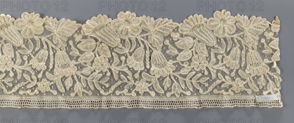 Lace fragment, floral ornaments; fragment of bobbin lace with relief edges made with a needle on a tulle background, featuring a motif of large flowers rosettes and bells and leaves; 19th century, Brussels; lace fabric, width 15 cm, length 110 cm., A wide piece of lace fabric with an overall cream color set against a gray background, featuring a dense floral and leafy pattern with several large scalloped flower motifs along the top edge and a group of bell-shaped blossoms and smaller flowers dispersed throughout; the lace has a fine netted ground with raised embroidered outlines and textured filling stitches creating petals, leaves, stems, and small round accents, and a horizontal decorative border along the bottom edge with open rectangular mesh and a narrow scalloped trim.
