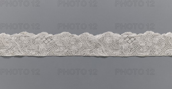 Lace fragment, floral ornaments, Binche lace, ca. mid-18th century, Binche place of origin ?, Henryków German: Heinrichau, lace, width 4.5 cm, length 102 cm, A horizontal strip of lace with a scalloped upper edge sits against a smooth background. The lace is light in color with an intricate floral and scroll pattern made of fine threads and openwork; areas of dense stitching contrast with delicate mesh. The scalloped top shows repeating rounded arches and tiny picot-like edges, while the lower edge is straighter and slightly uneven. Visible colors include off-white for the lace and gray for the background.