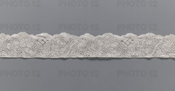 Lace fragment, floral ornaments, Binche lace, Henryków German: Heinrichau, lace, ca. mid-18th century, Binche place of origin?, lace, width 4.5 cm, length 102 cm, A horizontal strip of delicate white lace with a scalloped upper edge runs across the middle of the image against a smooth gray background; the lace shows an intricate floral and swirling pattern with areas of openwork and denser embroidered motifs, and the visible colors are white and gray.