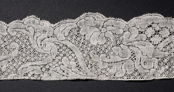 Lace fragment, floral ornaments, Binche lace, ca. mid-18th century, Binche place of origin?, lace, width 4.5 cm, length 102 cm, A length of delicate lace fabric laid horizontally against a dark background, showing an intricate openwork pattern with floral and scroll-like motifs; the lace appears off-white or cream with fine threadwork forming loops, scalloped edges, and areas of denser and more open stitching that create texture and shadow against the black or very dark gray surface beneath.