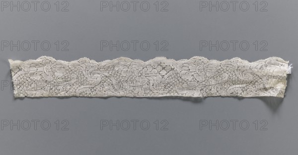 Lace fragment, bobbin lace, white, with a floral tendril ornament on a point de neige background, ca. mid-18th century, Binche place of origin?, fabrics, width 4.5 cm, length 32 cm, A long narrow strip of lace laid horizontally against a flat background, showing a scalloped top edge and a straight bottom edge with frayed ends; the lace has an open, intricate floral and scroll pattern with dense embroidered areas and mesh-like sections, appearing delicate and slightly irregular in shape. The visible colors are off-white and cream for the lace and a medium gray for the background.