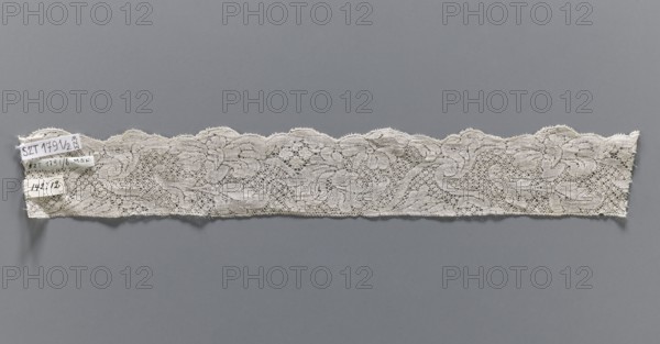Lace fragment, bobbin lace, white, with a floral tendril ornament on a point de neige background, ca. mid-18th century, Binche possible origin, fabrics, width 4.5 cm, length 32 cm., A long band of lace in off-white is displayed horizontally against a muted gray background, showing an intricate openwork pattern with floral and scroll-like motifs and a scalloped upper edge; the lace appears delicate with fine netting between the motifs and a slightly irregular lower edge, and there is a small cluster of fabric tags attached near one end in off-white and pale beige tones.