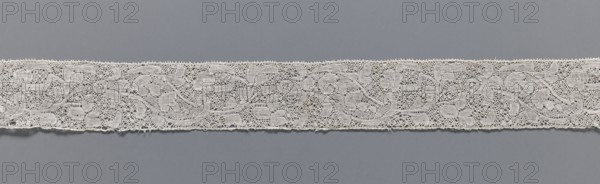 Lace fragment, lace, Binche lace, Henryków German: Heinrichau, clothing accessory, needle lace with point de neige background, straight edges, mid-18th century, Binche, width 3.5 cm, length 37 cm, A horizontal strip of delicate lace in an off-white or cream color runs across a smooth, muted gray background; the lace features a repeating floral and vine pattern with openwork areas and fine netting, scalloped tiny loops along the top and bottom edges, and a few small loose threads visible along the lower edge, while the background shows subtle variations of gray.