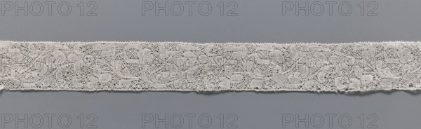 Lace fragment, lace, Binche lace, Henryków Heinrichau, clothing accessory, needle lace with point de neige background, straight edges, mid-18th century, Binche, width 3.5 cm, length 37 cm, A narrow band of delicate lace with an intricate floral and scroll pattern in off-white sits horizontally against a smooth gray background, showing a fine openwork texture with tiny loops and mesh-like areas, scalloped edges along the top and bottom, and a few loose threads along the lower edge.