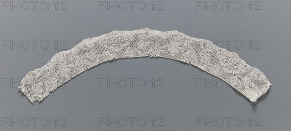 Lace fragment, Binche lace, slightly rounded in shape. The lower edge is slightly serrated, the upper one ends straight. Mid-18th century, Binche origin, lace, length 36 cm top edge: 30 cm; bottom edge: 36 cm, width 3.6 cm, A curved strip of lace in off-white spanning across a flat gray background, showing an openwork floral pattern with a mesh-like ground and scalloped upper edge; the lace has a few frayed ends and irregular edges, with visible texture, raised embroidered areas outlining floral motifs, and subtle shadows where it lifts from the gray surface.