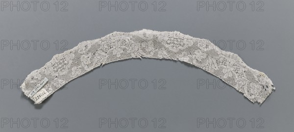 Lace fragment, mid-18th century, Binche lace, slightly rounded in shape; lower edge slightly serrated, upper edge straight; length 36 cm top edge 30 cm, bottom edge 36 cm, width 3.6 cm., A curved strip of lace with a floral and mesh pattern lies against a solid background; the lace is off-white with delicate scalloped edges and areas of fraying, and the background is a muted gray.