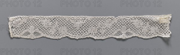 Lace fragment, mid-18th century. Binche lace, width 3.8 cm, length 30.5 cm. A tendril motif at the upper edge and ribbons with bows at the lower edge. Background in point de neige with drochelle bows in the loops; motif contours underlined with thread., A long narrow piece of lace laid flat against a solid gray background, showing an openwork pattern with floral and leaf motifs connected by a mesh of small hexagonal loops; the lace is primarily off-white or cream with subtle variations in shade, and its edges are slightly irregular with a few loose threads and small frayed areas.