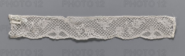 Lace fragment, repossession provenance noted, Binche lace, Henryków German: Heinrichau. Mid-18th century. A tendril motif at the upper edge and ribbons with bows at the lower edge. The background is a point de neige and in the loops are drochelle bows. The contours of the motifs are underlined with thread. Lace, width 3.8 cm, length 30.5 cm., A narrow strip of delicate lace with a repeating floral and geometric openwork pattern lies flat against a neutral background; the lace appears off-white to cream with a slightly textured surface and has scalloped edges and frayed ends, showing a group of small flower motifs connected by a network of fine threads and larger leaf-like shapes, with subtle variations in tone where threads overlap.