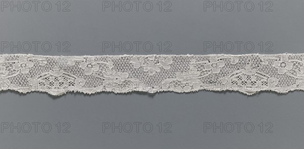 Lace fragment, Malines Mechelen lace, Henryków Heinrichau, varied background: point de neige type meshes and fanciful ones; contours of motifs highlighted with additional thread; mid-18th century; width 3 cm, length 50 cm., A horizontal strip of delicate lace with a floral and scroll pattern runs across a flat background; the lace is light-colored with intricate openwork, featuring a mix of solid embroidered flower shapes and fine netting with scalloped lower edges, and the background is a uniform cool gray.