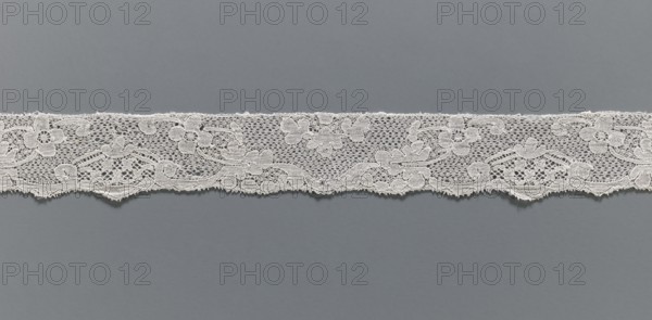 Lace fragment, reprovenance noted, lace, Malines Mechelen lace, Henryków Heinrichau, varied background: point de neige type meshes and fanciful ones; contours of motifs highlighted with additional thread; mid-18th century; width 3 cm, length 50 cm., A horizontal strip of delicate lace with a floral and scroll pattern lies across a flat background; the lace is light in color and intricately woven with openwork areas and solid embroidered motifs, showing scalloped lower edge and a straighter upper edge, while the background is a smooth medium gray.