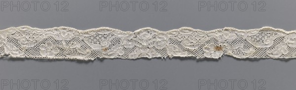 Lace fragment, mid-18th century, Mechelen Malines bobbin lace; fashion accessory. Motifs: floral sprigs, palmettes, leafy tendril, shells. Motif contours outlined with additional thread. Varied mesh background including point de neige. Gently serrated lower edge. Dimensions: width 3 cm, length 35 cm., A narrow strip of delicate lace with a repeating floral and scroll pattern lies horizontally against a smooth background; the lace shows open netting interspersed with solid embroidered motifs and scalloped edges, and there are small areas of fraying and a few darker discolored spots; visible colors include off-white and beige in the lace and a medium gray background.