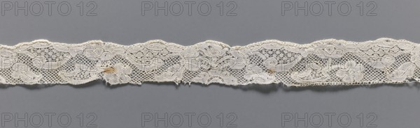 Lace fragment, repossession provenance; bobbin lace, Malines Mechelen style, mid-18th century; fashion accessory. Motifs: floral sprigs, palmettes, leafy tendril, shells; motif contours outlined with additional thread. Varied mesh background including point de neige. Gently serrated lower edge. Dimensions: width 3 cm, length 35 cm., A narrow strip of lace fabric runs horizontally across a gray background, showing an intricate floral and scroll pattern with open mesh areas and solid embroidered motifs; the lace appears off-white or cream in color with a few small tan discolorations and slightly frayed edges, and the background is a uniform medium gray.