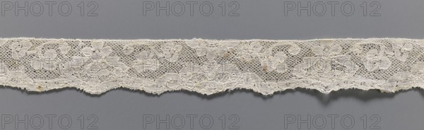 Lace fragment, lace, Malines Mechelen lace, motifs outlined with additional thread, Ißgrond background, lower edge slightly serrated, 2nd half of the 18th century, width 3.5 cm, length 37 cm, A horizontal strip of delicate lace with an irregular scalloped lower edge stretches across a gray background; the lace is a pale off-white or cream color with visible floral and looping mesh patterns and a few small darker spots and slight fraying along the edges.
