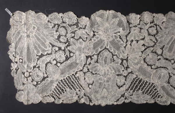 Lace fragment - ruffle, lace, plant patterns, floral patterns, palmettes ornament, clothing accessories, bobbin lace, Brussels lace. A ribbon runs across the entire width of the ruffle, dividing the surface into opposing triangles, fastened with either small palmettes similar to the fruit of a pineapple or larger leafy ones. The created triangular fields contain large motifs of leafy palmettes or palmettes on a pedestal. The backgrounds feature winding floral sprigs. Various mesh backgrounds with picots. The upper and lower edges are serrated to match the arrangement of the motifs. c. 1700, Brussels; lace; height 32 cm, width 230 cm, A length of delicate lace with an ornate floral and scrollwork pattern laid flat against a dark background; the lace is a pale off-white or cream color with a fine mesh filling and raised embroidered outlines forming large stylized flowers, leaves, circular motifs, and curved bands, along with scalloped edges and areas of openwork where the dark background...