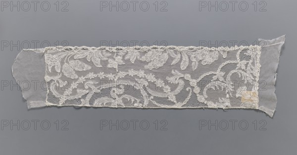 Lace fragment, A leafy tendril with fruit is arranged along the lower edge. At the upper edge, a garland of small flowers and a wide tendril with Rococo elements. The lower edge is slightly serrated, in line with the arrangement of the motifs. Mid-18th century, Mechelen. Lace, width 19 cm, length 67 cm., A rectangular piece of sheer fabric with lace overlay laid flat against a gray background, featuring ornate floral and scrolling motifs in an off-white or cream color; the lace has a scalloped top edge and a straight bottom edge, with a few torn or frayed extensions of plain, sheer fabric at both ends in a slightly grayer off-white tone, and a small patch of discoloration in a pale beige near one end.