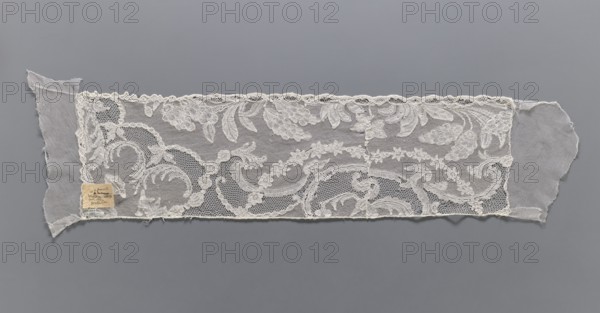 Lace fragment, A leafy tendril with fruit is arranged along the lower edge. At the upper edge, a garland of small flowers and a wide tendril with Rococo elements. The lower edge is slightly serrated, in line with the arrangement of the motifs. Mid-18th century, Mechelen. Lace, width 19 cm, length 67 cm., A long rectangular piece of delicate lace with floral and scrolling vine motifs laid flat on a smooth grey background, the lace appearing off-white to cream with areas of fine netting and scalloped edging, irregular frayed fabric extensions at each short end, and a small square of aged paper affixed near one corner showing a faded discolored tan color.