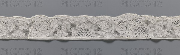 Lace fragment, lace, mesh background with rounded meshes. The lower edge is slightly serrated. The floral motifs on the edge are emphasized with thicker thread. 2nd half of the 18th century, Mechelen. Lace; width 4.2 cm, length 57 cm., A horizontal strip of lace with a scalloped top edge laid against a flat background; the lace shows an openwork floral and scroll pattern with areas of dense embroidery and net-like mesh, and a straight finished lower edge; visible colors include off-white in the lace and gray in the background.