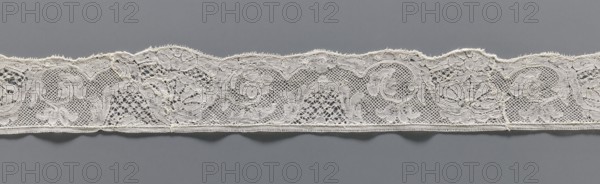 Lace fragment, lace, mesh background with rounded meshes. The lower edge is slightly serrated. The floral motifs on the edge are emphasized with thicker thread. 2nd half of the 18th century, Mechelen. Lace, width 4.2 cm, length 57 cm., A long narrow strip of delicate lace with a scalloped top edge and a straight bottom edge rests against a smooth background; the lace features a repeating floral and scrollwork pattern formed by open mesh areas and more densely stitched motifs, with visible loops and tiny decorative picots along the scalloped edge and a thin stitched border along the bottom; the lace appears off-white or cream against a medium gray background, and there are several areas where the threads form intricate nets and solid embroidered shapes.