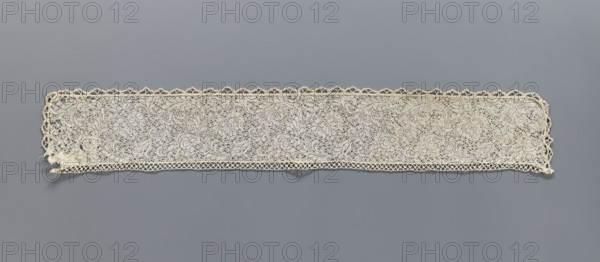 Fragment of bobbin lace with plant ornaments, 18th century, Flanders; width 7 cm, length 74 cm; maker, provenance: repossession., A long rectangular strip of lace displayed flat against a gray background, with a scalloped decorative edge along the top and a looped border along the bottom; the lace shows an intricate floral and scrollwork pattern with openwork areas and dense needlework, appearing delicate and slightly uneven at the ends; visible colors include off-white and cream in the lace and medium gray in the background.