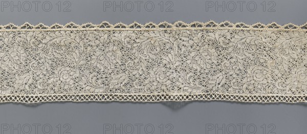 Fragment of bobbin lace with plant ornaments, 18th century, Flanders;, width 7 cm, length 74 cm., A length of delicate lace in off-white with an intricate openwork pattern sprawls horizontally across a neutral gray background, showing a floral and scroll motif composed of looping threads and airy mesh; the top edge features a scalloped decorative trim with small triangular openings, while the bottom edge has a narrow band of geometric openwork; the lace displays a soft cream color contrasted against the cool blue-gray backdrop.