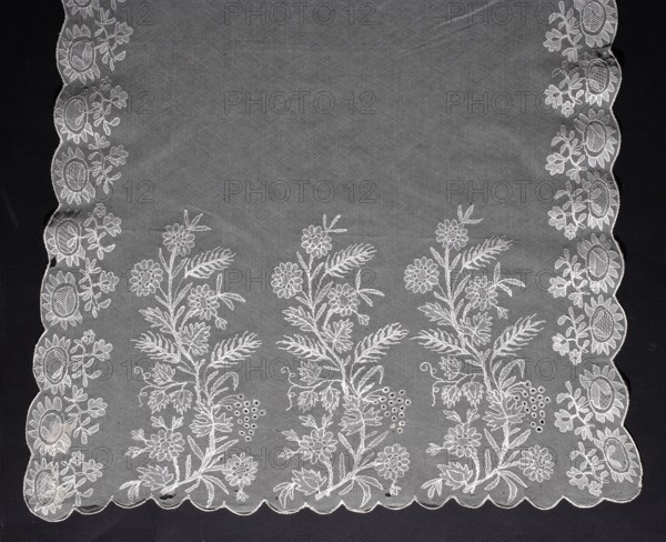 Shawl, floral ornaments; at each end are three flower sprigs with various plants such as ears of grain, vines, flowers and leaves; throws of flower sprigs around the longer edges; smooth interior; fashion accessory for women's clothing; 1820–1840; Europe; length 225 cm, width 59 cm., A rectangular embroidered textile with scalloped edges laid flat on a dark background, showing a delicate sheer fabric in a pale off-white color. Along the sides and bottom edge there are repeated floral embroidery motifs in the same pale off-white thread, forming vertical columns of flowers and leaves; the central area of the textile is mostly sheer with subtle texture of the netting. The side borders feature a repeating circular floral pattern with small leaves, while the bottom border displays several tall stems with clustered blossoms, leafy fronds, and small round berry-like motifs. The scalloped hem is outlined with denser stitching in the off-white color, and the background behind the textile appear...